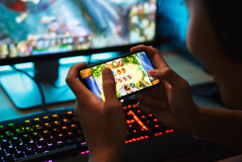 Portrait of teenage gamer boy playing video games on smartphone and computer in dark room